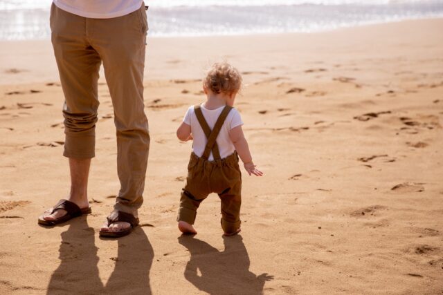 Anonymous dad looking after small kid carefully stepping on sandy beach in sunny day