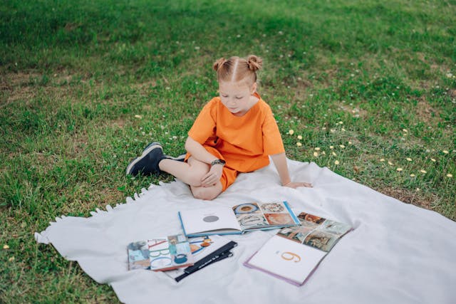 A young girl in an orange dress reading books outdoors on a picnic blanket.